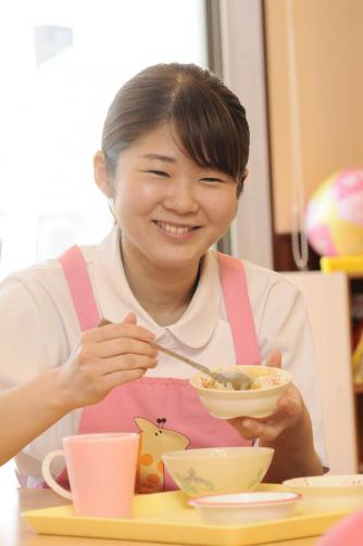 画像：ピンク色のエプロンをつけて、左手にご飯の入ったお茶碗を持ち右手に長いスプーンをもって笑っている新開さんの様子。