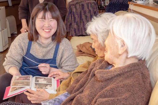 画像:施設内のソファーに座ってアルバムを開いている女性の利用者二人の隣で1枚の写真を左手に微笑んでいる渡邉さんの様子