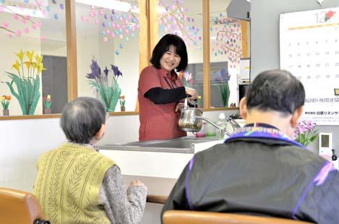 画像:手前に男性と女性の利用者の方が座っていて奥の流しでやかんを持った原田さんの様子。