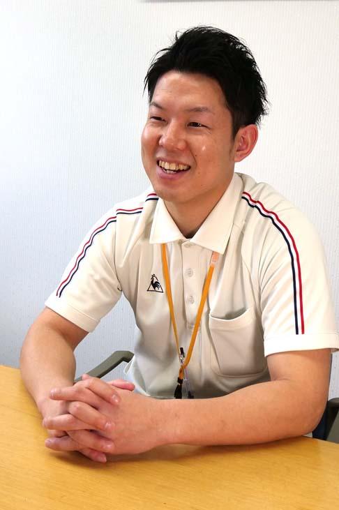 プロフィール写真:施設内の白い壁の部屋で両手をテーブルの上におき手を組んで明るくインタビューに答える脇田さんの様子