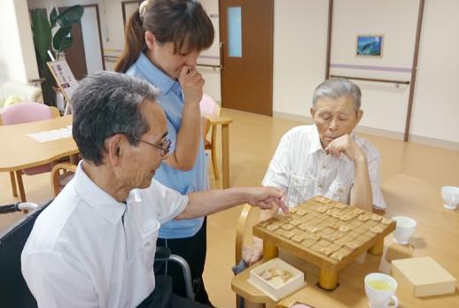 画像：施設内で２名の利用者が椅子に座って将棋を指しているところに介護者が間に立って将棋盤を眺めている様子