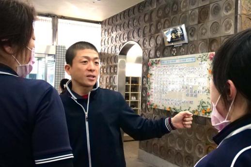 画像:施設内の利用者の一覧が貼ってあるボードの前に立ち二人の女性スタッフに対し何か話しをしている脇田さんの様子