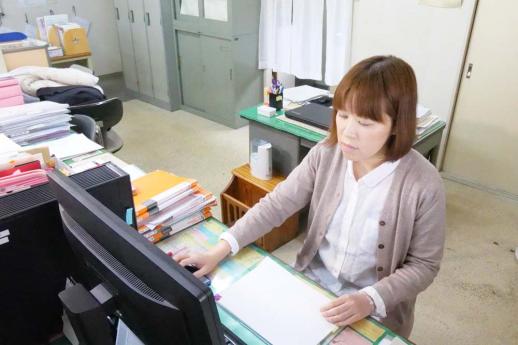 画像:施設内の事務室の机でパソコンに向かい左手に書類を持ち右手でマウスを操作している職員の様子