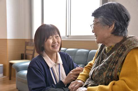 画像:ソファに座って笑顔で話をしている女性利用者の方と、しゃがんで笑顔で話を聞く岩永さんの様子。