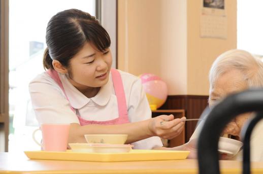 画像：ピンク色のエプロンをつけて女性の利用者さんの食事の介助をしている新開さんの様子。