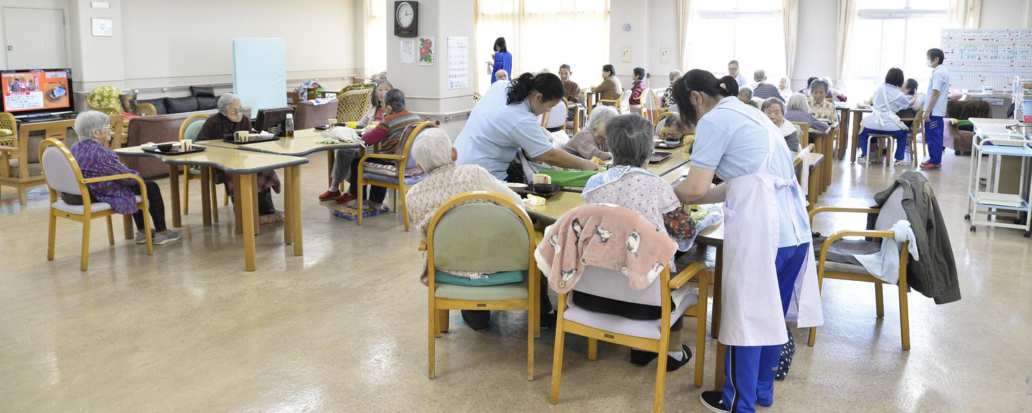 画像：施設内の食堂で複数のテーブルで食事している利用者と食事を介護している介護者の様子