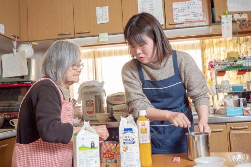 画像:施設内の台所で女性の利用者と一緒にカルピスや牛乳やオレンジジュースを使ってミックスジュースを作っている渡邉さんの様子