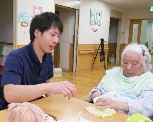 画像:施設の共有スペースの中で、左手で机の上のお絞りを触っている女性の利用者の方に隣に座って話しかけている江頭さんの様子