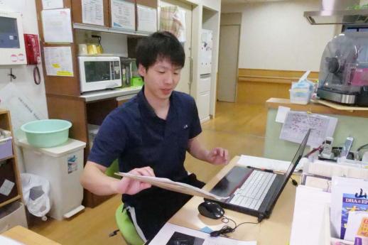 画像:施設のスタッフルームの机でノートパソコンを開き、大きな封戸を右手で持ち上げている江頭さんの様子