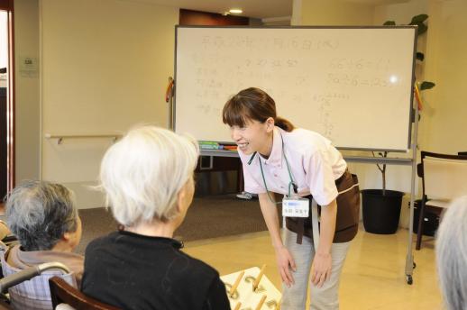 画像:ホワイトボードの前で中腰の姿勢で車椅子に座っている女性の利用者さんに目線を合わせ笑いながら話している永田さんの様子。
