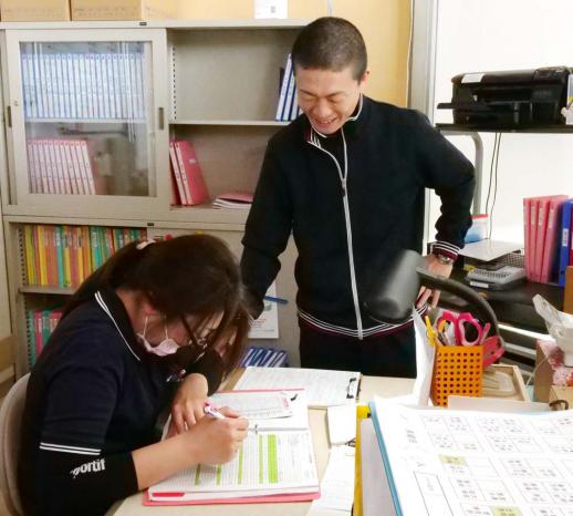 画像:施設内の後ろのキャビネットに書類が入っている事務所の中で、事務机に座って書類を記載しているスタッフの脇に立ち指導をしている脇田さんの様子