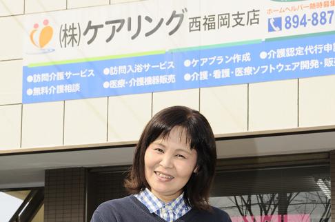 画像:タイル調の壁に社名の看板が貼ってあり、その前で微笑んでいる中島さんの様子。