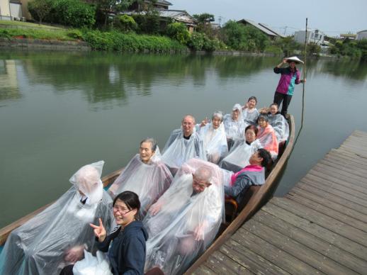 画像:川下りの船に船着き場の近くで2列に並んだ11人の白い雨合羽を着た利用者と一名の立っている船頭さんの一緒に船に乗っている松門さんの様子