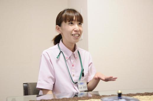 画像:白い壁の部屋で、ピンク色のポロシャツを着て左手を軽く上げでいる永田さんの様子。