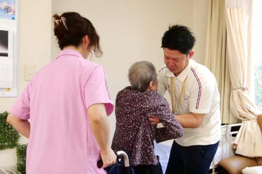 画像:車いすを押す女性スタッフと車いすから利用者の両脇を抱えて移動を介護している脇田さんの様子