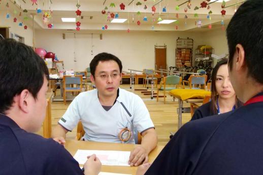 画像:施設のリハビリ施設の中で志田さん含め4名スタッフがテーブルを囲み座りミーティングをしている様子