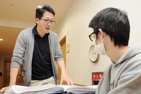 画像:男性スタッフの前で机の上にファイルを広げ立った状態で話している川畑さんの様子。