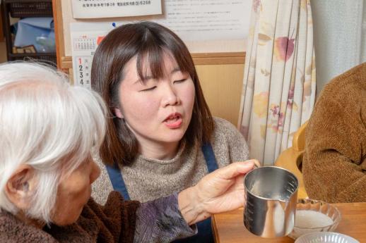 画像:施設内で利用者2名の間にしゃがみ、利用者1名の方が左手に計量カップを持っているそばから声をかけている渡邉さんの様子