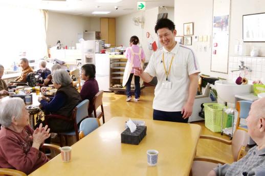 画像:施設内の食堂で湯呑が二つ置かれているテーブルを挟んで座っている利用者に対して、立って左手で利用者を指しながら話をしている脇田さんの様子