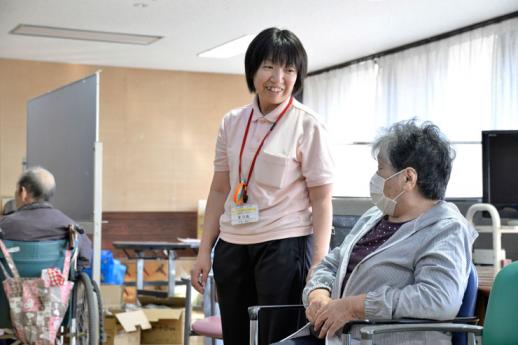 画像:施設内の食堂の一角で椅子に座っているマスク姿の女性利用者と立ったまま笑顔で会話する松尾さんの様子