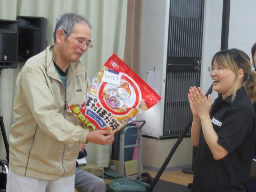 画像:施設内の共有スペースで男性利用者の方がぽたぽた焼きの大きな袋を持っているそばで手を叩いて喜んでいる松門さんの様子