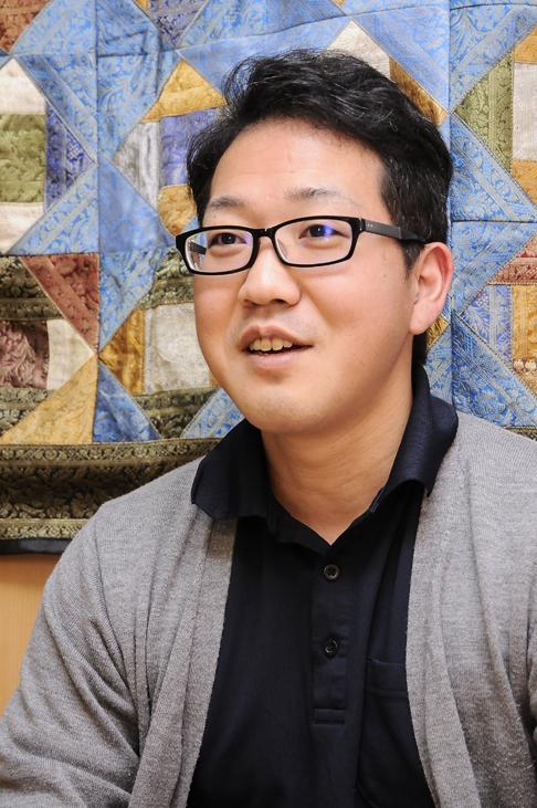 プロフィール写真:パッチワークの作品がかかっている壁の前でインタビューに答える川畑さんの様子。