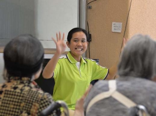 画像：施設内で利用者の前で両手をあげ、にこやかに体操しているエルニタ・プランシスカ・プルバさんの様子