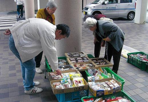 画像:施設のピロティに複数のかごの上に段ボールサービス箱に移動販売品が並べ、その品物を4名の利用者が選んでいる様子